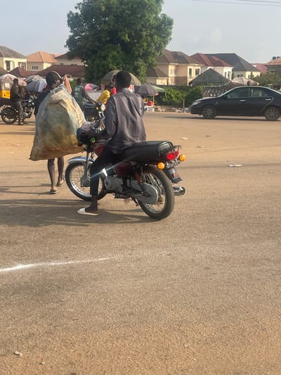 In Galadimawa Village, Nigeria, a street scene unfolds with a focus on a motorcycle and its occupants. A man, dressed in a gray shirt and dark pants, is seated on the motorcycle, which is positioned on a paved road. He is wearing white shoes. Another individual, also wearing dark clothing and a cap, is seen carrying a large, light-colored sack. This sack appears to be made of woven material and has red and blue lettering on it, with the visible text reading "AVIA PREMIUM RICE". The motorcycle itself has a red tail light and orange turn signals, and appears to be loaded with cargo, possibly within a black container behind the seat.
In the background, several houses with distinctive architectural styles, featuring peaked roofs, are visible. Green trees dot the landscape, suggesting a warm climate. The road is asphalt, marked with a white line on the right side. The lighting suggests it is daytime, with clear skies. The overall impression is one of everyday activity and commerce in a Nigerian village setting.