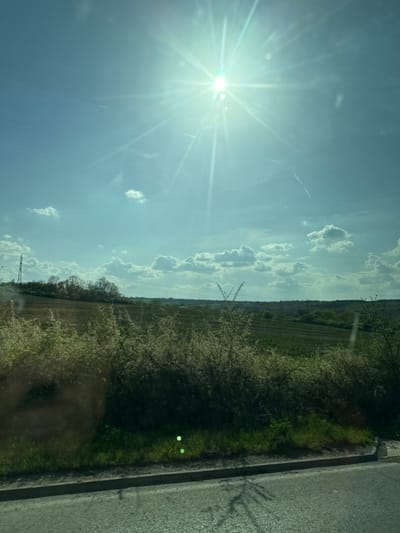 The image presents a bright, sunlit rural landscape, likely captured from a moving vehicle. The primary subject is an expansive sky, dominated by a brilliant, low-angle sun in the upper left, causing pronounced lens flare with radial light streaks and circular halos. The sky is a clear, light blue with scattered white cumulus clouds, some appearing wispy, others more defined, and faint contrails are visible.

A dark asphalt road occupies the right side of the image, suggesting the perspective is from alongside or looking out from a vehicle. A gray curb or edge separates the road from a vibrant green grassy verge. This verge and the middle ground are densely lined with trees and bushes, many adorned with white blossoms, indicating spring or early summer. Further into the distance, agricultural fields stretch towards a distant horizon, with faint power lines or pylons visible against the sky. Two distinct, bright green circular reflections or lens anomalies are present on the green verge in the lower right.

The time of day is clearly daytime, likely late morning or early afternoon due to the bright sun. The weather is clear and sunny. The setting is a rural or countryside environment, consistent with agricultural land and roadways. There is no visible text in the image. The original file is rotated 90 degrees clockwise from a standard landscape orientation.