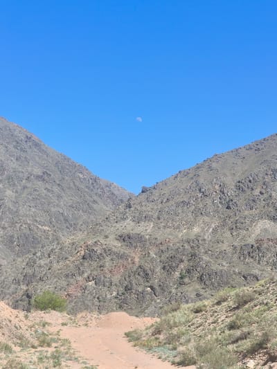 A natural landscape photograph depicts a valley in the region of Krasnyy Most, Kyrgyzstan, during daylight. The scene is dominated by two large, converging mountainsides, which are sparsely vegetated and covered in a mix of brown, gray, and green rock formations. A worn, earthen trail leads up the center of the frame into the valley between the mountains. The sky is a clear blue, and a slightly visible moon is positioned in the upper center of the frame. The landscape is devoid of any visible human presence or structures apart from the trail itself.