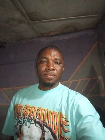 A man with a shaved head and short beard wears a light blue t-shirt with a large, colorful graphic. He stands against a textured wall. The lighting is dim. Two markings are visible on the left side of his face, near his eye and cheek. The ceiling above appears to be metallic or industrial.