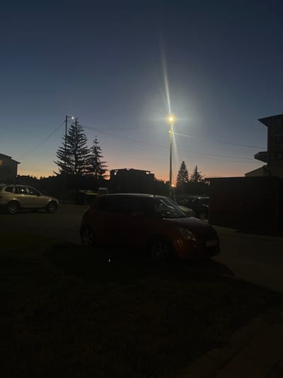 In Samokov, Bulgaria, at dusk, a streetlamp illuminates the scene. In the foreground, a red car is parked on the left side of the road. Beyond it, a silver SUV is parked on the right. In the background, silhouetted against the twilight sky, are trees and buildings. The sky transitions from deep blue at the top to hues of orange and pink near the horizon, indicating the setting sun. The overall impression is a quiet residential street at the end of the day.