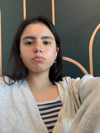 A woman is depicted in a selfie, oriented at an angle. Her hair is dark and she has an earbud in her left ear secured with a safety pin. She wears a striped top and a grey cardigan. The background consists of a dark green wall with curved, light brown lines. There are no clear indicators of the city of Austin.