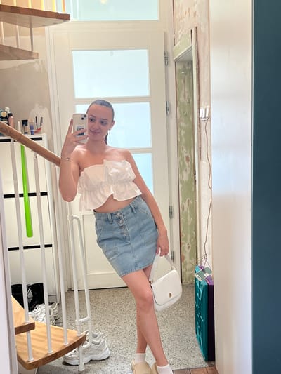 A fair-skinned female takes a selfie in what appears to be an indoor environment, possibly a private residence. She holds a smartphone with her left hand, which is equipped with a white case, positioned to capture her image. She is wearing a white, ruffled top, a light blue denim skirt with button details, white socks, and light-colored shoes. A small, white handbag hangs off of her shoulder.
In the background, there is a door with multiple glass panels and a partially unfinished ceiling area. A white clothes rack is visible with various items, including shoes and clothing. Various cleaning supplies and bottles rest on a nearby shelf. The color palette primarily consists of white, blue, brown, and green. The location in the city of Saarbrücken, Germany, cannot be determined from this image.