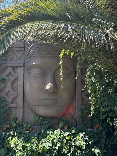The image depicts a large, stone relief of a serene face, possibly a Buddha or a deity, embedded in a wall. The relief is adorned with intricate patterns on either side of the face. Lush green foliage, including palm fronds and vines, partially obscures the stone structure, creating a natural, somewhat overgrown aesthetic. Red accents are visible among the foliage, possibly flowers or decorative lights. The overall scene suggests an outdoor setting, possibly a garden or a decorative wall feature in a public space. The lighting indicates it is daytime, with sunlight filtering through the leaves. The location of this artwork is in Asprovalta, Greece.