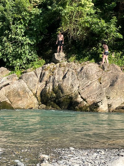 In Plastunka, Russia, two shirtless individuals, a male and a female, are positioned on a large, layered rock formation adjacent to a body of water. The male is standing on a higher section of the rock, facing away from the camera, wearing dark swim trunks. The female is on a lower section of the rock, facing the camera, also wearing dark swim trunks. The rock formation is covered in patches of moss and vegetation, with dense green foliage forming a backdrop behind them. The water in the foreground is clear and turquoise, with ripples indicating movement. The riverbed is visible in places, covered with various sized pebbles and stones. The scene suggests an outdoor recreational setting, possibly for swimming or cliff jumping, given the natural environment and the attire of the individuals.