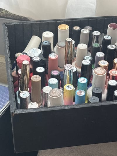 A black storage container, possibly a drawer or box, is filled with numerous small, cylindrical bottles, likely nail or gel polishes, in Jesús del Monte, Mexico. The bottles are arranged horizontally, creating a dense, somewhat disorganized collection. Their caps display a wide array of colors and finishes, including metallic, glitter, solid, and translucent shades of red, pink, gold, silver, black, white, cream, blue, teal, and purple.
Many caps feature circular labels with numbers and some text. Clearly visible numbers include 043, 394, D6, 56, 044, 004, 709, 001, E143, 85, 52, 14, and 10. Text visible on specific caps includes "BASE FRANCESA" on a pink octagonal cap and "RED FRANCESA" on another. The background shows hints of a white fabric on the left and a darker, textured surface on the right. No people are present, and the bright, even lighting suggests daytime.