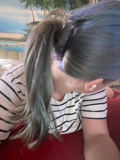 The media file is a close-up, portrait-style photograph of a person's head and upper torso. The person has long, dark hair with subtle streaks of blue and purple. The hair is partially pulled back in a ponytail, revealing a small, white earbud in their right ear. They are wearing a white and black striped long-sleeved shirt. A silver chain necklace is visible over the shirt. The background appears to be a wall mural depicting a beach scene with palm trees, sand, and the ocean under a blue sky, suggesting an indoor setting or a decorative element. The person is lying down, with their head resting on a red surface. The lighting is soft, and there are no strong shadows, indicating it could be daytime or well-lit indoor conditions. The overall mood is relaxed and casual. No visible text is present in the image. The provided location context is Nova Praha, Ukraine.