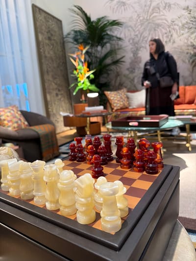 The image depicts a well-appointed indoor space, likely a hotel lobby or a upscale lounge, situated in Rho, Italy. The primary focus is a large chess set positioned on a dark, rectangular table or stand. The chess pieces are distinctly chunky and translucent, with one set in amber and the opposing set in a creamy white color, arranged on a brown and tan checkered board.
In the blurred background, a person, appearing to be a woman with dark hair and dark clothing, is standing or moving near a patterned wall. The room is furnished with comfortable seating, including armchairs or sofas, accessorized with decorative, patterned cushions. A large potted plant with palm-like leaves and another plant featuring bright orange and green Bird of Paradise flowers add greenery to the space. A framed artwork or mirror is visible on the wall behind some furniture. The wall itself has a subtle, light-colored pattern. Partial window blinds are visible at the bottom left, suggesting natural daylight illuminates the scene. No specific activity is occurring, and there is no visible text. The overall atmosphere appears calm and inviting.