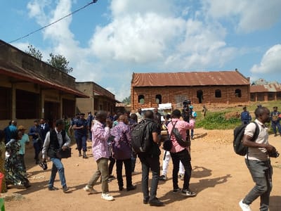 The image shows a group of people gathered in front of a building in Beni, Democratic Republic of Congo (latitude: 0.497, longitude: 29.485). There are about 20 people in the image, some standing, some walking, and some talking to each other. The majority of the people are men, and they appear to be dressed in casual clothing. There are also a few women in the image, one of whom is holding a small child. The building in the background is made of brick and has a rusty metal roof. The building appears to be unfinished, as there is no window glass in the windows. The ground is dirt and there are some trees and shrubs in the background. The sky is blue with a few white clouds. In the center of the image, a man with a black backpack appears to be filming the scene with a video camera, while a man wearing a pink shirt stands with a microphone in his hand. Other people are holding their cell phones.