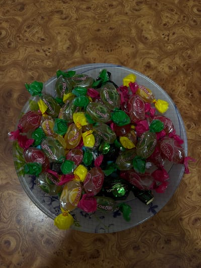 A close-up overhead shot shows a clear glass bowl filled with colorful, individually wrapped candies. The bowl sits on a polished wooden surface with a distinct, swirling grain pattern. The candies are a mix of fruit flavors, indicated by their wrappers: green for apple, red for cherry, and yellow for lemon. Some wrappers are partially transparent, revealing the candy inside. The wooden surface is warm-toned, with rich brown and amber hues, and appears to be a tabletop or countertop. The lighting is soft and diffused, casting minimal shadows. There is no indication of any specific location in the city of Mahilyow, Belarus, or any people present in the image. The scene is static and appears to be a still life arrangement of candy.