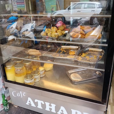 The image shows a display case from "Cafe Atharva", filled with assorted food items and jars of ingredients. On the top shelf are fried snacks, including what appear to be vada. The next shelf down contains trays with remnants of what seem to be fried items, alongside a tray of what may be papads. The bottom shelf holds several jars filled with yellow and white substances, likely ghee and lassi, and a tray with a single item. Outside the case are a few bottles, including a green glass bottle with a straw. Reflections in the glass reveal scooters, a white car, and other street elements. The location is in Oxel, India.