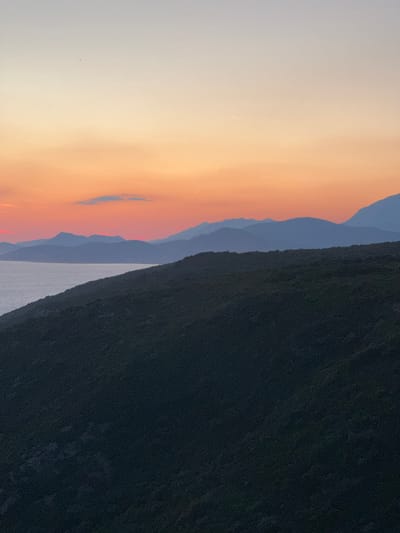 A panoramic view of a tranquil sunset over the Adriatic Sea near Glavatičići, Montenegro, is depicted. The sky is a gradient of soft hues, transitioning from a pale peach at the horizon to a serene lavender at the zenith. Distant, silhouetted mountain ranges, layered in shades of blue and purple, recede into the fading light. In the foreground, a dark, densely vegetated hillside slopes down towards the calm, grey sea. The overall impression is one of natural beauty and peaceful solitude.