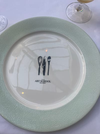An overhead shot captures a white dining plate with a textured, pale mint green rim, set on a plain white tablecloth. Centered on the white interior of the plate are three silhouetted kitchen utensils: a whisk on the left, a chef's knife in the middle, and a spoon on the right. Beneath these implements, the text "THE ART SCHOOL" is printed in a larger font, with "PAUL ASKEW" appearing in a smaller font directly below it. Partial views of two stemmed glass tumblers are visible on the tablecloth: one with an etched pattern on the upper left, and another containing a yellowish liquid on the upper right. The plate's surface exhibits slight reflections of ambient light sources. This setting is indicative of The Art School restaurant, operated by Paul Askew, located in Liverpool, United Kingdom. No people or active interactions are depicted.
