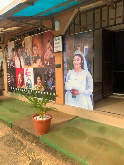 The image shows a display of photographs outside a building, likely a photography studio. The photographs feature various individuals, including what appears to be a bride in a wedding dress at the top of the display. Below this, there are several other portrait shots of people, some of whom seem to be children and adults. One photograph prominently displays the text "HAPPY BIRTHDAY" with a balloon and a baby. Another smaller sign reads "Book YOUR SESSIONS NOW." The overall setting appears to be outdoors, with some architectural elements suggesting a covered walkway or storefront. A potted plant with green fronds sits in front of the display, adding a touch of nature to the scene. The ground is a mixture of concrete and a green surface, possibly artificial turf. The photographs are mounted on a wall or board, suggesting they are advertisements for the studio's services. The lighting suggests it is daytime. The visible text indicates the location is Sabojibi, Nigeria.