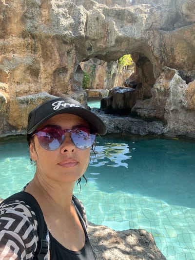 A woman with dark hair and sunglasses is taking a selfie in a swimming pool. She is wearing a black visor and a black and white patterned cover-up over a black swimsuit. The pool is turquoise and appears to be in a tropical setting with rock formations and greenery in the background. The location is the Ayana Resort and Spa in Bali, Indonesia.