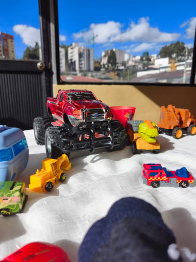 This is a close-up, eye-level shot of various toy vehicles arranged on a white, textured surface, likely a tablecloth or sheet, indoors. The background, visible through a window or opening, depicts a city skyline with buildings and a bright blue sky with clouds. The city appears to be Quito, Ecuador, based on the architectural style of the buildings and the hilly terrain.
In the foreground, a large, metallic red toy Ford F-350 truck with oversized tires is prominently displayed. To its left is a light blue toy vehicle with windows, possibly a train car or RV. To the right of the red truck, a yellow toy excavator with large black wheels sits. Further to the right, a toy truck with a dinosaur head as its cargo is visible. Behind these, an orange toy construction vehicle with a crane is positioned. In the lower right corner, a red and blue toy fire truck is placed. Partially obscuring the foreground is the out-of-focus back of a dark blue knitted object, likely a hat or part of a garment. A red toy car is also visible at the bottom left, mostly out of focus. The lighting suggests a bright, sunny day, casting shadows from the toys.
