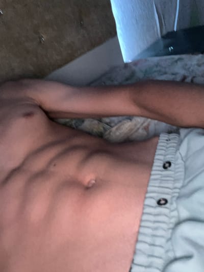 The image shows a close-up of a person's torso, focusing on their abdomen and arm. The person appears to be lying down, with their arm resting behind their head. The abdominal muscles are clearly defined, indicating a toned physique. They are wearing light-colored shorts with a striped pattern and buttons. The background is blurred, but appears to be a bedroom setting with a bed and a hint of natural light coming from a window. There are no discernible features that would suggest the location in Ognianovo, Bulgaria.