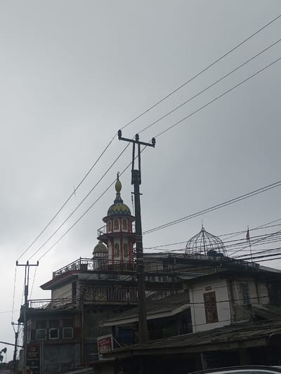 An overcast sky looms over a multi-storied structure capped with Islamic architectural elements. A red and white minaret and a wire-framed dome accent the building. Utility poles and a web of wires intersect the view, obscuring the detailed architecture. Below, weathered rooftops and advertisements are visible. The building blends residential and commercial aspects.