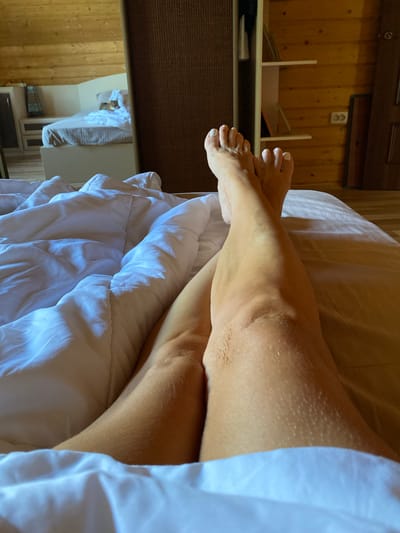 A close-up, point-of-view shot shows a person's bare legs and feet crossed on a bed with white bedding. The legs are tanned, and the toenails are painted white. The background consists of a wooden-paneled room, possibly a bedroom in a cabin or dacha. A portion of a mirror reflects a bed with a cat on it, and wooden shelves with frames and other items. The room appears to be located in a rural setting, possibly in or near the village of Gabishevo, Russia, given the architectural style and mention of the location. The lighting suggests it's daytime, with natural light entering the room.
