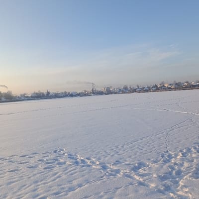 A snow-covered landscape under a pale blue sky. A town or village is visible in the distance, partially obscured by mist or haze. A prominent chimney or industrial structure emits smoke. Footprints and tracks mark the expanse of the snow-covered ground, suggesting activity or movement across the frozen surface. The sun's rays create shadows and highlights, adding depth and texture to the snow.