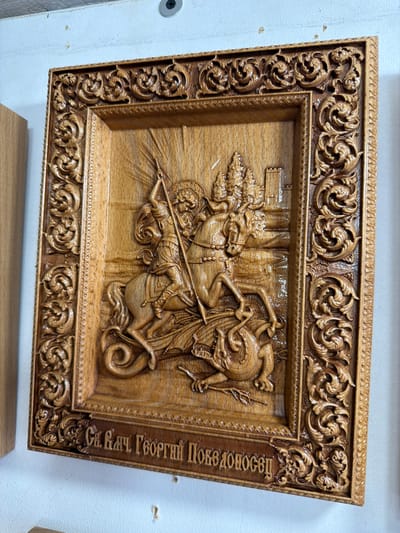 The image shows a carved wooden icon depicting Saint George slaying a dragon. The icon is framed with intricate scrollwork. Below the carving, there is an inscription in Cyrillic script. The icon is mounted on a white wall, with a portion of a wooden object visible to the left. The background appears to be an indoor setting with a white ceiling. No specific location within the city of Yalta, Ukraine can be determined from the image.