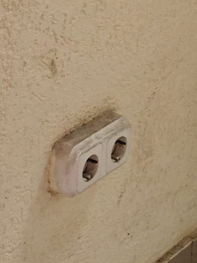The media file shows a close-up, eye-level shot of a double electrical socket mounted on a textured, light-colored wall. The socket is white and rectangular with two oval openings. It appears to be somewhat dusty and aged. The wall itself has a rough, stippled finish and is a neutral, creamy beige color. There are no people or active events depicted in the image. The focus is solely on the electrical outlet and the surrounding wall texture. The lighting suggests it could be indoors, possibly in a residential or public building, and there are no external environmental cues. No text is visible in the image. Given the location context of Svetlahorsk, Belarus, this is likely an interior view of a building in that city, depicting a common household or public utility fixture.