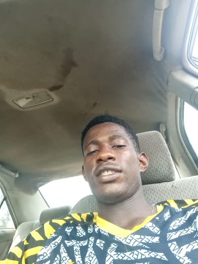 A selfie inside a car. A young Black man with short hair and a yellow and white patterned shirt smiles slightly. The car's interior is visible, including the seat headrest, window, and ceiling. Stains mark the car's ceiling above the person, and a light fixture is present near the roof. Outside the window, hints of greenery and daylight can be seen. The angle is from slightly below the person's face.