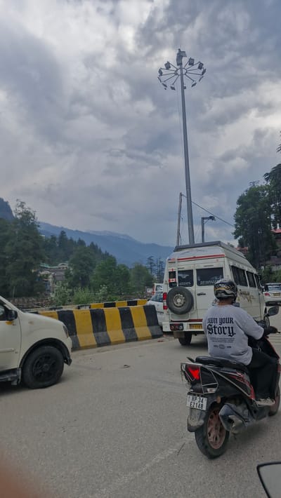 In Manali, India, under a cloudy sky, a traffic scene unfolds on a paved road. A person on a scooter, wearing a grey t-shirt with the text "Own your Story.", is in the foreground, facing away from the camera. Behind the scooter, a white van with a spare tire mounted on the rear is visible, as is another white car. A yellow and black striped barrier runs along the side of the road. In the background, lush green trees and mountains, some with snow-capped peaks, create a scenic backdrop. A tall pole with multiple lights is also present in the scene.