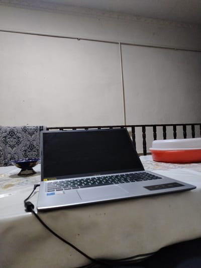 This is a photograph of a laptop computer placed on a table, likely indoors. The laptop is silver and open, with its screen facing the viewer. The keyboard is visible, and a black power cord is plugged into the laptop and trails off to the left. In the background, there is a white wall with a pipe running horizontally across it. To the left of the laptop, there is a patterned fabric draped over what appears to be a sofa or bench, and a decorative bowl sits on the table. To the right of the laptop, a dark wooden railing separates the table from a red and white plastic container. The lighting suggests it might be evening or an indoor setting with artificial light. The presence of specific Uzbek cultural patterns on the fabric and the distinct style of the railing and container could suggest a location in Uzbekistan, such as Nukus.
