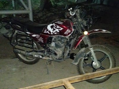 A red dirt bike with a white wolf decal is parked on a paved surface in Danghara, Tajikistan. The motorcycle is oriented diagonally, with the front wheel on the right side of the image and the rear wheel on the left. It appears to be a night scene, with artificial lighting from the motorcycle's headlight and possibly other sources. The bike is somewhat dirty, suggesting it has been ridden off-road. In the background, there are some blurred out trees and a fence or railing. A wooden plank is visible in the foreground, partially obscuring the front wheel of the motorcycle.