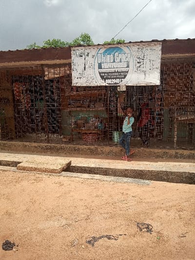 A young girl, wearing a blue t-shirt, blue jeans, and pink sandals, is standing in front of a shop with a black metal gate in Ota, Nigeria, located at latitude: 6.689, longitude: 3.178. The sign above the shop reads "Divine Grace Ventures, Wholesales & Retails For all your daily needs at the best prices 08023940172". There is a drain in the foreground, and the ground is dirt. There is a small, brown, wooden crate on the left side of the shop. Two black plastic bags are on the ground in the foreground. There are trees in the background. The sky is cloudy. The shop appears to be closed.