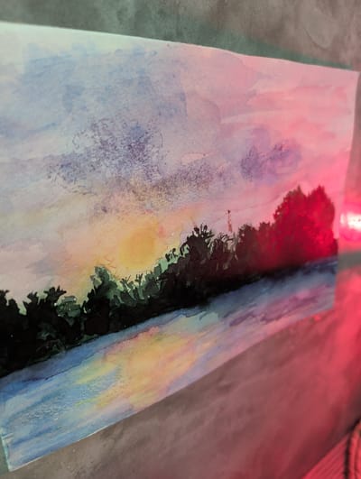 A watercolor painting depicting a landscape is displayed on a gray surface under red lighting. The painting features a sky with pink, purple, yellow, and blue hues, transitioning into darker shades near a line of trees. The trees are depicted as dark silhouettes, and in the center, there's a suggestion of sunlight. The painting's composition creates a sense of depth and perspective. The painting is angled slightly to the right. The scene depicts a nature scene in Ko Pha-ngan, Thailand.