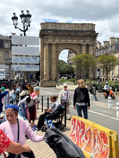 The image depicts a lively street scene in Bordeaux, France, near the Porte Cailhau. In the foreground, an elderly woman wearing a pink knitted sweater and a blue headscarf is visible. She is interacting with another person whose arm is extended, holding a dark garment. To the right, a woman in a wheelchair, wearing a white t-shirt with a black and white patterned design, is seated. She has a cup in her hand. Behind them, other individuals are seen amidst what appears to be a street market or gathering, with clothing displayed on the ground.

Dominating the background is the grand arch of Porte Cailhau, a historic monument in Bordeaux. The sky is cloudy, suggesting overcast weather. The street is paved, and there are traffic lights and bollards visible. A person on an orange scooter is crossing the street in the middle ground. Several street lamps and signage are also present in the scene. The overall atmosphere appears to be one of casual public activity on a city street.