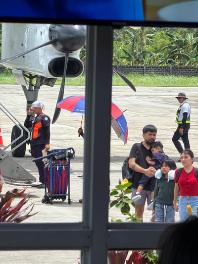 From an indoor vantage point, the scene unfolds at the Del Carmen airport in the Philippines. The foreground is dominated by a window frame. Outside, a plane and ground crew are visible. A man in a reflective vest stands with equipment, while another individual holds a red and blue umbrella. Adjacent to them, a small plane has a visible propeller. On the right side of the frame, a family, including a man carrying two young children, stands on the tarmac. There is a woman with them wearing a red sweater. An airport worker in a reflective vest stands nearby. Tropical vegetation lines the background of the airport.