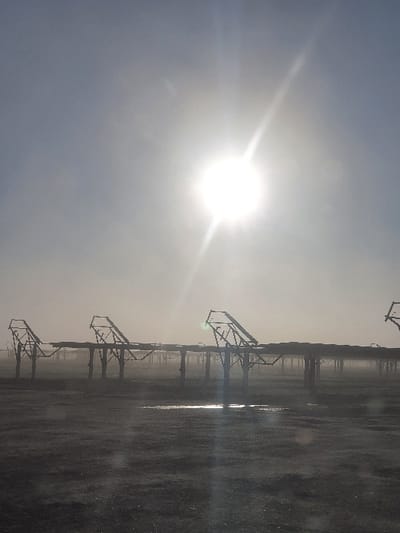 A bright sun shines in a hazy sky, partially obscured by clouds. Silhouetted against the glare are structures resembling irrigation systems or industrial equipment. They appear in a linear formation along a gray, reflective surface, possibly wet ground or standing water, creating mirroring effects. The scene is dominated by the sun's luminescence, resulting in low contrast and obscuring details in the foreground. The overall impression is desolate and slightly ethereal.