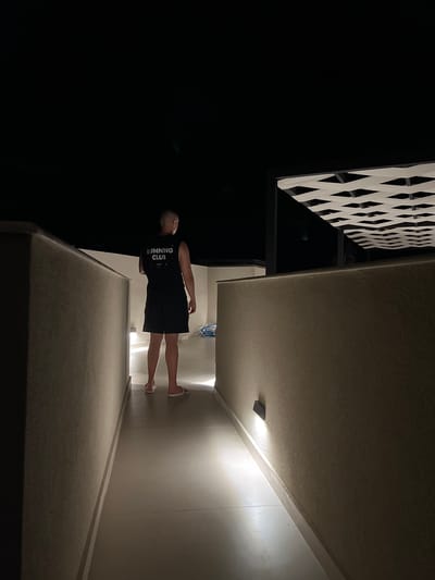 A person stands with their back to the camera, walking down a dimly lit, outdoor walkway at night. They are wearing a black sleeveless shirt with "RUNNING CLUB" in white lettering on the back, black shorts, and white flip-flops. The walkway is bordered by textured walls on both sides, with upward-facing lights illuminating the path. Above, a white lattice-like structure with dark supports is visible against the black night sky. The overall scene suggests an outdoor architectural space in Sithonia Municipal Unit, Greece.