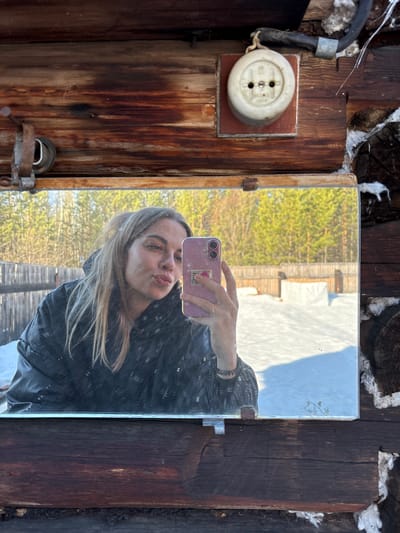 A person is taking a selfie in a mirror attached to a weathered wooden wall. The reflection shows a blonde person wearing a black jacket, holding a pink iPhone. Outside the reflection, the wooden wall has a vintage electrical outlet. The setting appears rustic, perhaps a dacha or shed. There is white material stuffed in the spaces between the wall logs. The exact location within Zheleznodorozhnyi, Russia cannot be determined from the image.