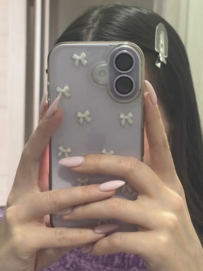 The scene is located in Novosaratovka, Russia. The image displays a close-up, reflected view of an individual, likely female, from the upper torso to just above the head. The subject's long, dark hair is parted down the center, with a clear or light-colored, arched hair clip positioned on the right side of the head. Both hands are holding a smartphone vertically, with fingers displaying long, pointed nails painted in a light pink or nude shade. The smartphone is enclosed in a translucent purple case featuring multiple small, white, three-dimensional bow embellishments. The rear of the smartphone shows a camera module with two vertically stacked dark lenses. A textured purple garment, presumably worn by the individual, is visible in the lower portion of the image. The background is out of focus and indistinct, consistent with a reflection in an indoor setting.