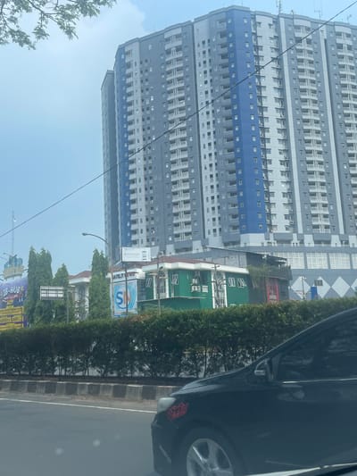The image captures a daytime scene of a residential building in Tangerang, Indonesia. A tall, multi-story building with a grey and blue facade dominates the background. In the foreground, a black car is partially visible, driving on a road. The scene is bordered by lush green hedges and trees, with a light-colored sky overhead. A street light is visible in the middle ground, along with several smaller buildings. Billboards and signage are present, indicating a commercial area. The overall impression is a typical urban landscape.