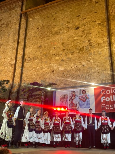 This media file shows a group of people in traditional folk costumes, likely participating in a festival or performance. The scene appears to be outdoors, as indicated by the stone wall in the background, which has an aged and textured appearance. Red and white lights illuminate the subjects, creating a vibrant and festive atmosphere, suggesting the event is taking place in the evening or at night.
The individuals are dressed in elaborate costumes featuring white blouses with intricate embroidery, colorful woven vests, and long white skirts with decorative trim. Some of the women wear headscarves. The men are also in traditional attire, including vests and dark pants. They are arranged in a line, with some holding hands, suggesting they are either performing a dance or posing for a photograph. Their expressions appear focused and engaged.
A banner in the background displays text, part of which is legible as "Cale Folk Fest." This, along with the "Calella, Spain" location context, confirms the event is a folk festival in Calella. The banner also features images of people in traditional clothing, further reinforcing the cultural theme. The setting is a performance area, possibly a street or a square, with the stone wall providing a historical backdrop. The lighting, including red stage lights, suggests a theatrical or public event. The overall impression is one of cultural celebration and community engagement.