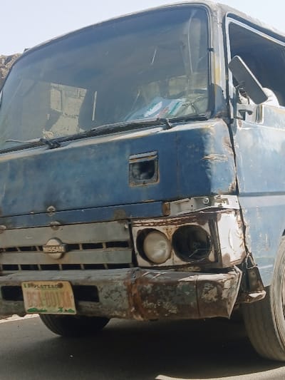 The image displays a severely dilapidated light blue, front-facing Nissan truck, stationary on a paved surface under a bright sky. The vehicle exhibits extensive corrosion and structural damage, particularly on its front fascia. The metallic front bumper is heavily rusted, dented, and missing sections, revealing underlying components. A "NISSAN" emblem is centered on the horizontal grille bars, which also show signs of wear. The passenger-side (viewer's left) headlight is intact but aged, while the driver-side (viewer's right) headlight assembly is missing its light unit, leaving an empty, discolored housing.

A yellow and green license plate, bearing "PLATEAU" at the top and "BSA-801XA" below, is affixed to the lower front bumper, indicating registration in Plateau State, Nigeria. The requested location of Masvingo, Zimbabwe, is not supported by the visible vehicle registration.

The truck's paint is faded, scratched, and peeling, revealing rust spots and underlying material on various body panels, including the hood, fender, and cab. The windshield appears dirty or scratched, with some papers or objects visible on the dashboard inside the cab. A rectangular side mirror is attached to the driver's side door frame. No individuals are visible in the scene. The immediate background shows a partial view of a light-colored structure to the left.