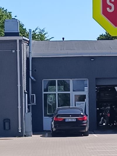 A black car with the license plate "UL-PB10" is parked in front of a gray building with a large open garage door. The building appears to be a service station or car repair shop. A sign with a red and yellow hexagonal shape and the letter "S" is visible in the upper right corner of the image. Trees and a blue sky are visible in the background. The scene is outdoors, on a paved surface. The location is in the city of Grobiņa, Latvia.