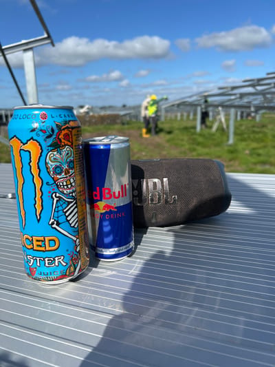 The scene is outdoors, apparently in a construction area, with a partially visible metal frame structure in the background. The sky is blue with some clouds. Close-up, there is a dark-colored JBL speaker, a can of Red Bull, and a can of Monster Juice drink on a ribbed metal surface. In the background, there are figures of people who appear to be working near the metal frame. The location could be inferred from the prompt to be in or around Grange Ville, Ireland.