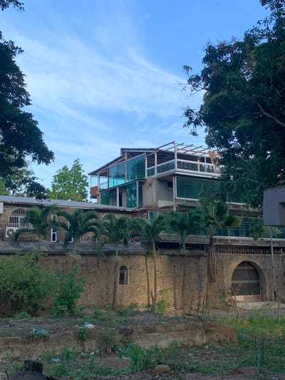 The image shows a multi-story building under construction, or possibly undergoing renovation, in El Valle del Espíritu Santo, Venezuela. The building has a modern design with large glass windows and balconies. It is surrounded by lush green trees and vegetation. A stone wall with an arched entrance and gate is in the foreground, separating the building from a somewhat unkempt area with scattered debris and foliage. The sky is a clear blue with some wispy clouds. The scene appears to be captured during daylight, likely in the late afternoon or early morning, given the soft lighting. There are no people visible in the image.