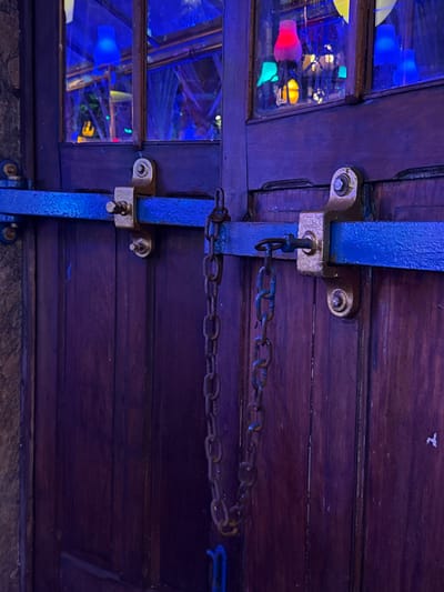 This image depicts the exterior of a dark-stained wooden double door, illuminated by a deep blue and purple ambient light, located in Rio de Janeiro, Brazil. A horizontal blue metal security bar spans the upper portion of the doors, affixed by two visible gold-toned metal brackets bolted into the wood. A rusted metal chain hangs vertically from the right side of this blue bar, connected by a metal ring. Above the security bar, the upper panels of the door feature multi-paned windows, through which an interior space is visible. This interior is illuminated by various colored light sources, appearing as distinct lamps or lanterns, including prominent blue, pink, yellow, and green lights. A textured, possibly stone, wall is visible on the far left edge of the frame. No human subjects or active events are present.
