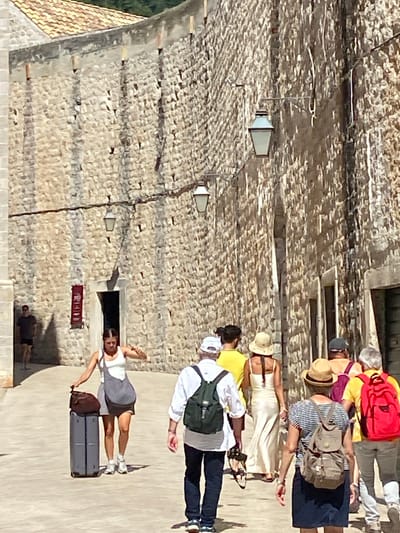 The image shows a view of a street within the city walls of Dubrovnik, Croatia. The street is paved with light-colored stone, and on either side are very tall, ancient stone walls. The walls are constructed from irregular, rough-hewn stones and are topped with a weathered, darker coping. Along the right-hand wall, several old-fashioned street lamps are mounted. In the distance, a glimpse of a tiled roof and some greenery suggests the presence of buildings and vegetation beyond the walls.
Several people are walking along the street. In the foreground, a woman with dark hair, wearing a white tank top and grey shorts, is pulling a large, grey suitcase with a brown duffel bag on top. She is wearing white sneakers. To her right, a person wearing a white shirt, dark pants, and a green backpack is walking away from the camera. To the right of them, another person in a straw hat, a white dress, and sandals is walking ahead. Further back, a group of people, including one in a yellow shirt and another in an orange shirt, are also walking away. On the far left of the image, a man in a dark t-shirt and shorts is standing near an arched doorway, which has a red banner hanging next to it. The scene is illuminated by bright sunlight, casting strong shadows.