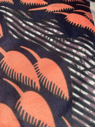 The image shows a close-up of fabric with a pattern featuring abstract orange shapes with serrated edges against a dark blue background. Interspersed are diagonal lines of white and purple. The fabric appears to be of a synthetic material, possibly polyester, with a visible weave. The pattern is abstract and does not represent any recognizable objects or scenes. There are no discernible landmarks or environmental features that would allow for the identification of its location within Jimeta, Nigeria. The image is a still, and there are no people or actions depicted.
