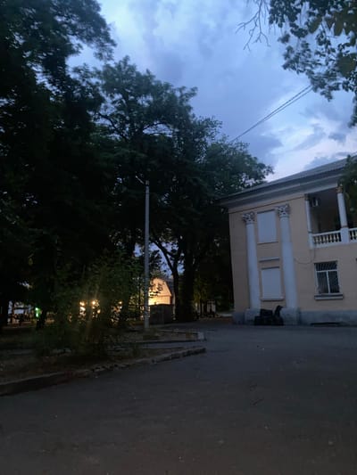 The image shows a building with a light yellow facade and white columns, likely in Voznesensk, Ukraine, as suggested by the query. The building has a covered balcony with a white railing and a tiled roof. There are several large trees with dark green foliage in the foreground and to the left, obscuring parts of the scene. The sky is overcast with dark clouds, suggesting it might be dusk or a stormy day. A paved area, possibly a street or courtyard, is visible in the foreground. There are a few dark, indistinct objects placed near the base of the building. The lighting is dim, with some artificial lights glowing faintly from behind the trees on the left. The overall impression is a quiet, somewhat somber outdoor scene in an urban or suburban setting.