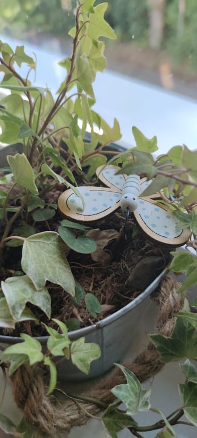 A close-up, eye-level shot captures a decorative wooden butterfly nestled among the leaves of an ivy plant in a metallic pot. The butterfly, painted white with blue polka dots and grey stripes on its body, sits with its wings spread. The pot is wrapped with coarse brown rope. The background consists of a blurry windowpane with reflections of the outdoor environment, suggesting the plant is indoors near a window, possibly in a home or establishment in Liebenburg, Germany. The lighting is natural and diffused, highlighting the textures of the leaves and the decorative elements. There are no people or visible actions taking place.