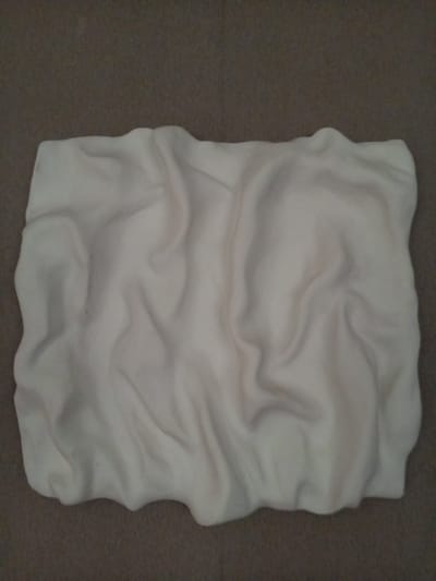 The image is a top-down, close-up shot of a folded, cream-colored fabric, likely a blanket or throw, placed on a dark, textured surface resembling carpet. The fabric is soft and has numerous wrinkles and folds, creating shadows and highlights that give it a somewhat three-dimensional appearance. There are no people or animals visible in the image. The background is uniform and out of focus. The image does not contain any discernible elements that would allow for the identification of its location within the city of Vilyuysk, Russia, or any other specific geographical location.